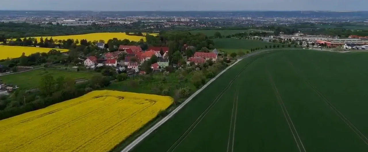 Drohnenaufnahme über Bannewitz: ein alter Dorfkern mit roten Dächern zwischen Rapsfeldern und Äckern, am Horizont der freie Blick bis zur Stadt