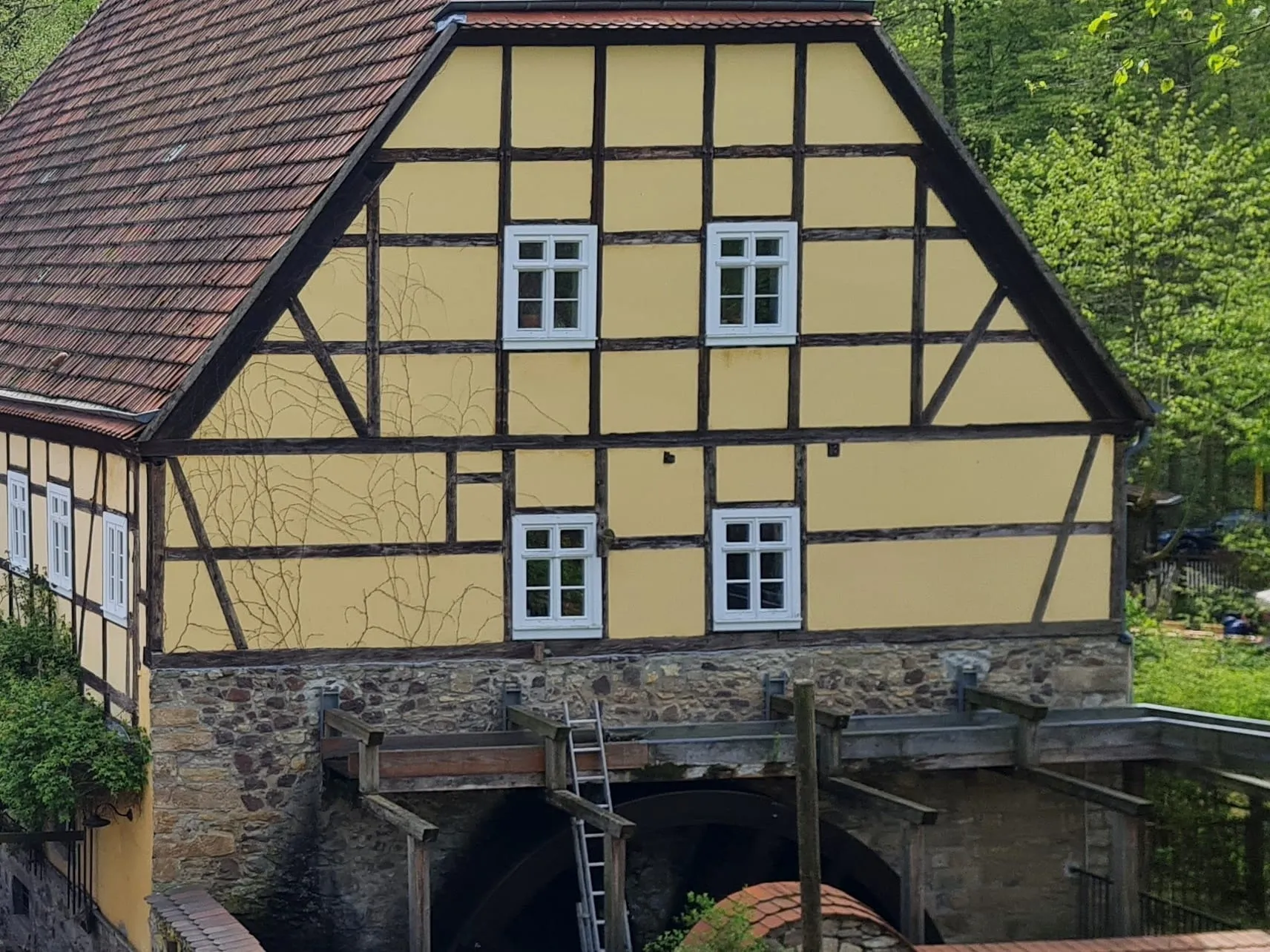 Foto der Zschonermühle im Zschonergrund: gelb gefasstes Fachwerkhaus mit rotem Walmdach über dem gemauerten Mühlgraben, ringsum der Frühlingswald des Tals.