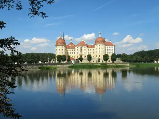 Schloss Moritzburg, das barocke Wasserschloss bei Dresden, spiegelt sich mit seinen vier Rundtuermen und roten Kuppeln im ruhigen Schlossteich unter blauem Himmel